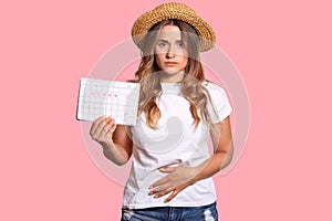 Displeased European young female feels pain and spasm during menstruation period, holds calendar, keeps hands on bottom of belly,