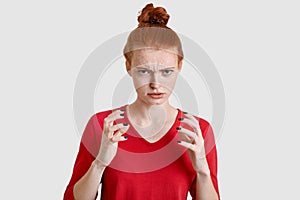 Displeased foxy young woman has freckled skin, keeps hands in angry gesture, looks with anger, wears red clothes, isolated over