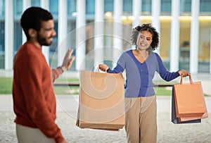 Displeased Husband Looking At Happy Wife With Shopper Bags Outdoors