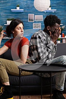 Displeased interracial couple sitting on sofa in silence