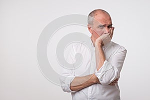 Displeased mature man in white shirt covering his face with hand over gray background.