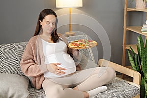 Displeased pregnant woman eating pizza alone at home sitting on sofa in living room overeating fast food during pregnancy touching