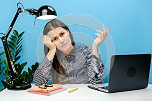 Displeased sad employee young woman 20s in office sit at desk work on laptop pc computer, posing isolated over blue color wall in