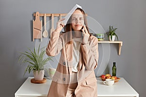 Displeased sad woman with brown hair wearing beige jacket standing in home kitchen interior having conversation with boss makes