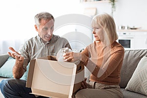 Displeased Senior Couple Unpacking Cardboard Box At Home