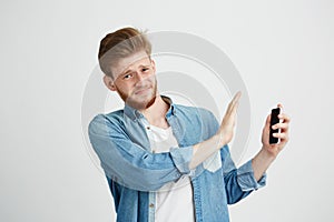 Displeased young handsome man showing stop sign to phone in another hand looking at camera resisting over white