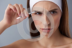 Displeased young lady plucking eyebrows with tweezers