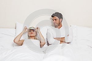 Displeased young man looking at woman wearing eye mask in bed