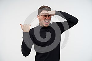 Displeased young short haired bearded male in glasses closing his nose with fingers while showing disgust and pointing back with