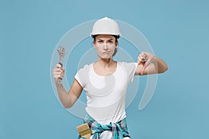 Displeased young woman in protective helmet hardhat hold adjustable wrench isolated on blue background. Instruments