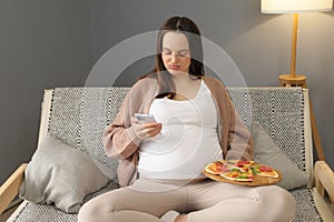 Displeased young woman sitting on sofa holding plate with pizza using smartphone browsing internet web pages while eating