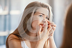 Displeased young woman squeezing acne on her nose while looking in mirror indoors