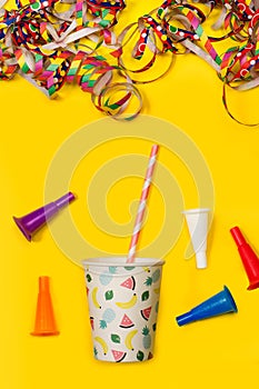 A disposable cup with a striped straw, serpentines and toy cornets on a yellow background