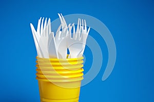 Disposable plastic tableware on blue background. White forks and knives, a yellow cups