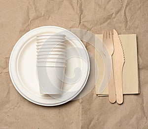 Disposable round white paper plates and cups on brown paper background, top view