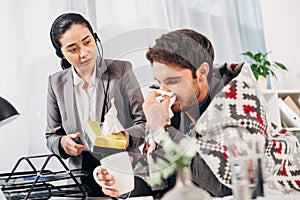 Disquieted call center operator giving napkins to sick coworker with blanket and cup