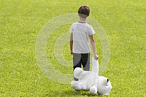 Dissapointed young boy is holding a teddy bear and standing on the meadow. Child looking down. Back view. Sadness, fear