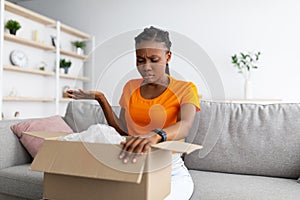 Dissatisfied black woman unpacking cardboard box, receiving damaged item at home. Bad delivery service, online shopping