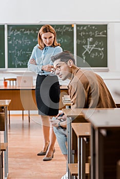 Dissatisfied female teacher with arms crossed looking at male student cheating during exam