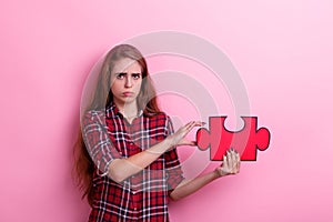 Dissatisfied girl holds a large puzzle her hand. Pink background.