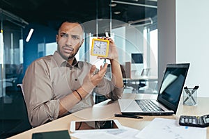 Dissatisfied hispanic man looking at camera and showing clock, businessman frustrated waiting for colleague to be late