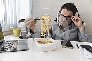 dissatisfied man eating instant noodles while working with laptop in office. Lunch at the office. tasteless junk food