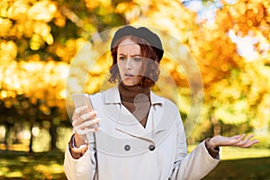 Dissatisfied sad confused caucasian millennial woman with red hair in raincoat, looks at phone, gesticulate