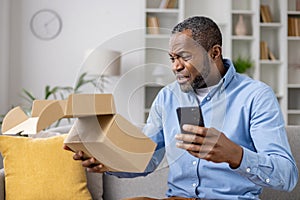 Dissatisfied sad online buyer, man received wrong damaged parcel, african american man sitting at home on sofa in living