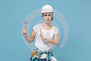 Dissatisfied woman in protective helmet hardhat showing stop gesture with palm to adjustable wrench isolated on blue