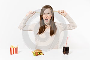 Dissatisfied woman at table with several pieces of potatoes, french fries, burger, cola in bottle isolated on white