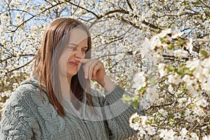 Dissatisfied young adult woman having allergy symptoms from blooming tree pollen in spring rubbing her nose posing near cherry