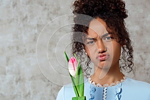 Dissatisfied Young African girl in a blue dress with a Tulip on a gray background. The concept of women`s day and spring