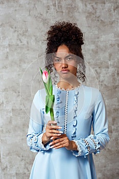 Dissatisfied Young African girl in a blue dress with a Tulip on a gray background. The concept of women`s day and spring