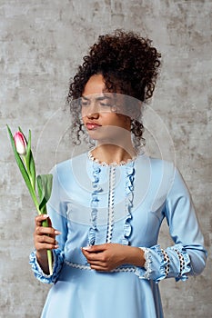 Dissatisfied Young African girl in a blue dress with a Tulip on a gray background. The concept of women`s day and spring