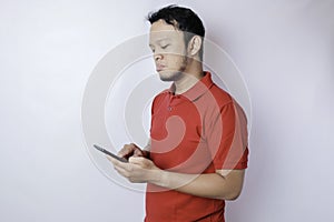 A dissatisfied young Asian man looks disgruntled wearing red t-shirt irritated face expressions holding his phone