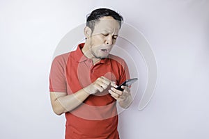 A dissatisfied young Asian man looks disgruntled wearing red t-shirt irritated face expressions holding his phone