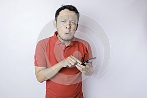 A dissatisfied young Asian man looks disgruntled wearing red t-shirt irritated face expressions holding his phone