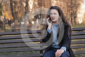 Dissatisfied young woman sits in park on bench and talks on phone and gesticulating. Girl in autumn park