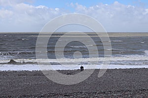 In the distance, a couple stand at the waters edge and watch the waves and the sea at Bossington Beach in Somerset, England