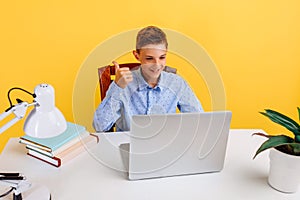 Distance education for children. schoolboy sitting at a table, learning by video link, happy and showing a thumbs up on a yellow