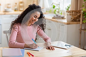 Distance Education. Woman using digital tablet in kitchen and writing in notebook