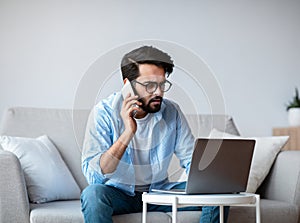 Busy Eastern Freelancer Guy Talking On Phone And Using Laptop At Home