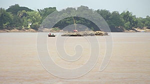 Distance view of a small motorized dugout canoe pulling behind a large heap of bamboo poles down the river 