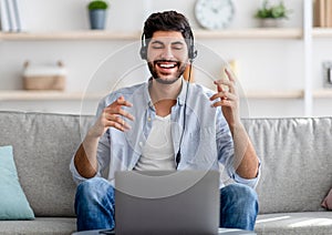 Distant communication concept. Happy millennial arab guy in headset talking and gesturing to laptop camera