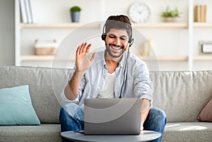 Distant communication concept. Smiling arab man in headset waving hand at laptop camera, making video call