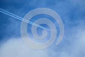 Distant passenger jet plane flying on high altitude through white clouds on blue sky leaving white smoke trace of