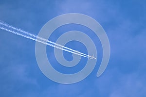 Distant passenger jet plane flying on high altitude through white clouds on blue sky leaving white smoke trace of