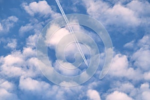 Distant passenger jet plane flying on high altitude through white clouds on blue sky leaving white smoke trace of