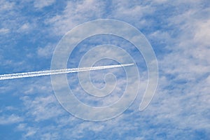 Distant passenger jet plane flying on high altitude through white clouds on blue sky leaving white smoke trace of