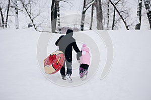 Distant photo of female kid and father with sled going up hill to slide down in snow in forest. Astonishing background